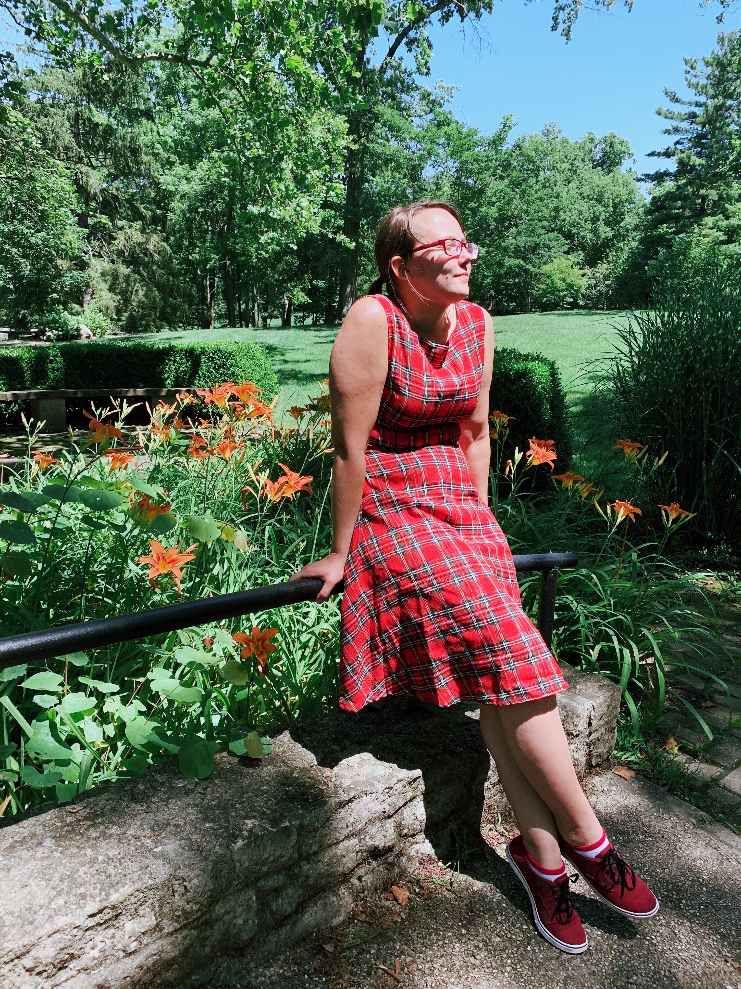 picture of the writer leaning against a railing outside in a red plaid dress. orange tiger lilies are behind the rail