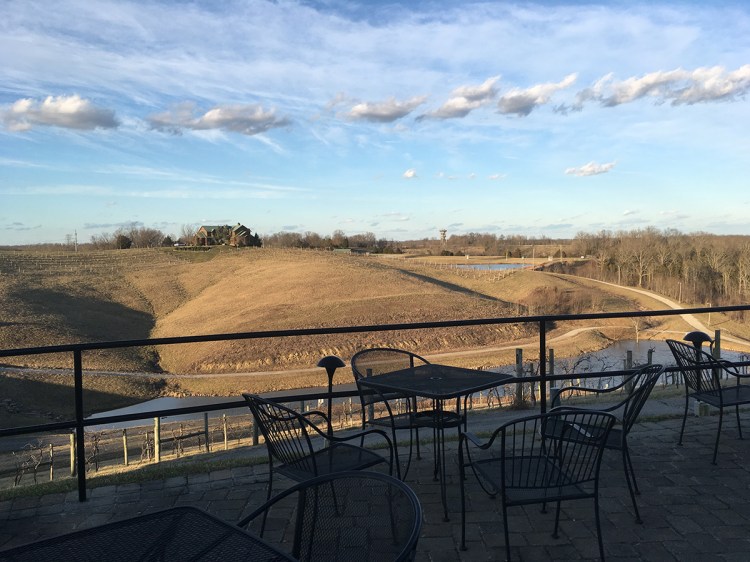 The view of a hillside in the summer from the porch of a winery. In the foreground, metal chairs and tables can be seen. 