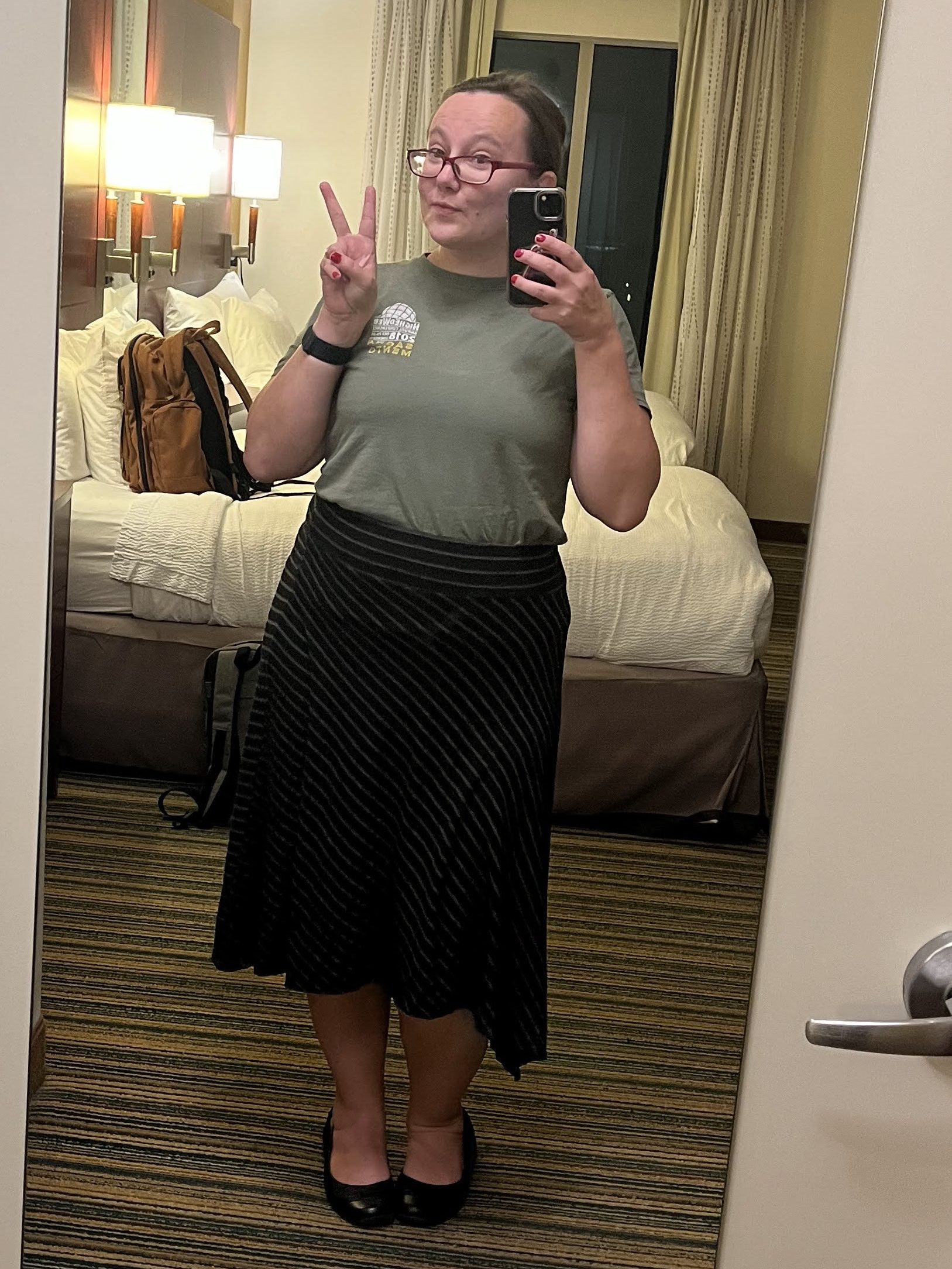 Liz, a glasses-wearing, brown-haired, white, female-presenting person, gives the "peace" sign in a mirror selfie in a hotel room in Arkansas.