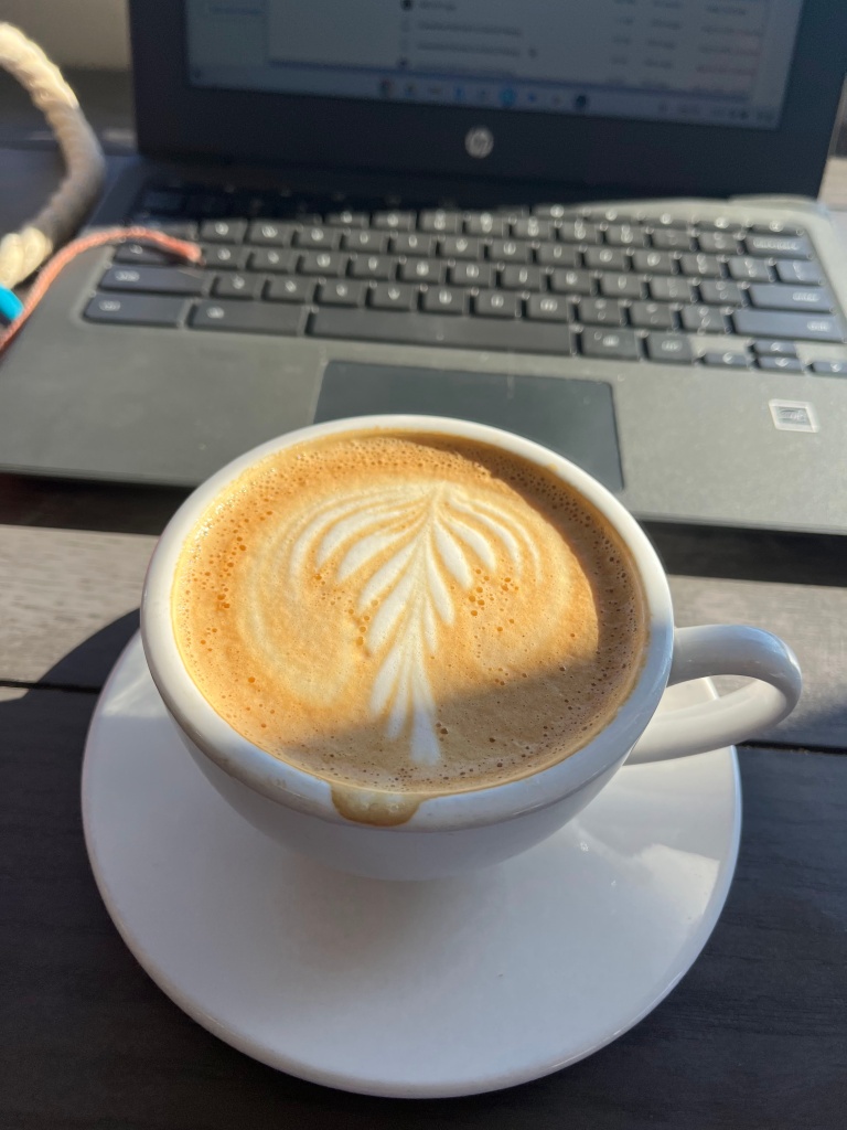 A cappuccino with latte art resembling a leaf, in front of a laptop