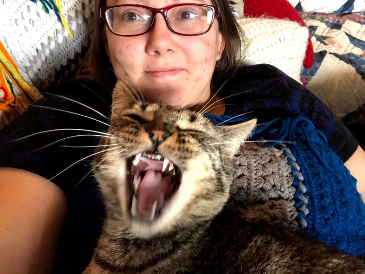 Liz, a feminine-presenting person wearing red-rimmed glasses, holds a yawning tabby cat.