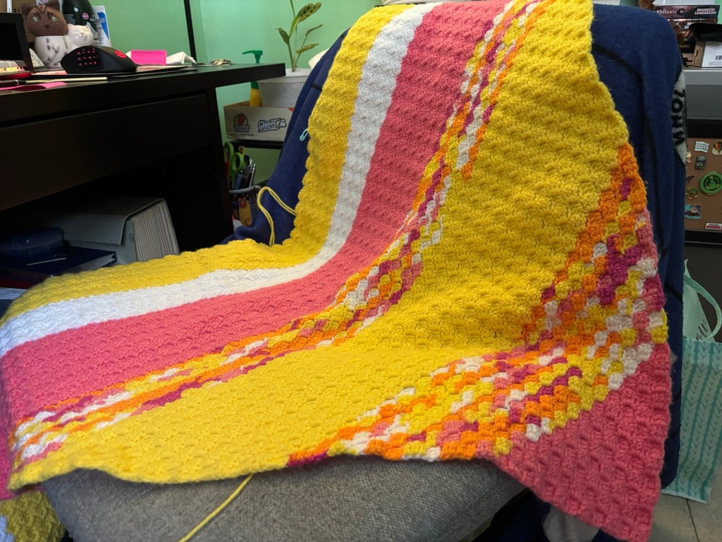 Half of an unfinished blanket draped over a chair. The color scheme is pink, yellow, white, and variegated of all the above
