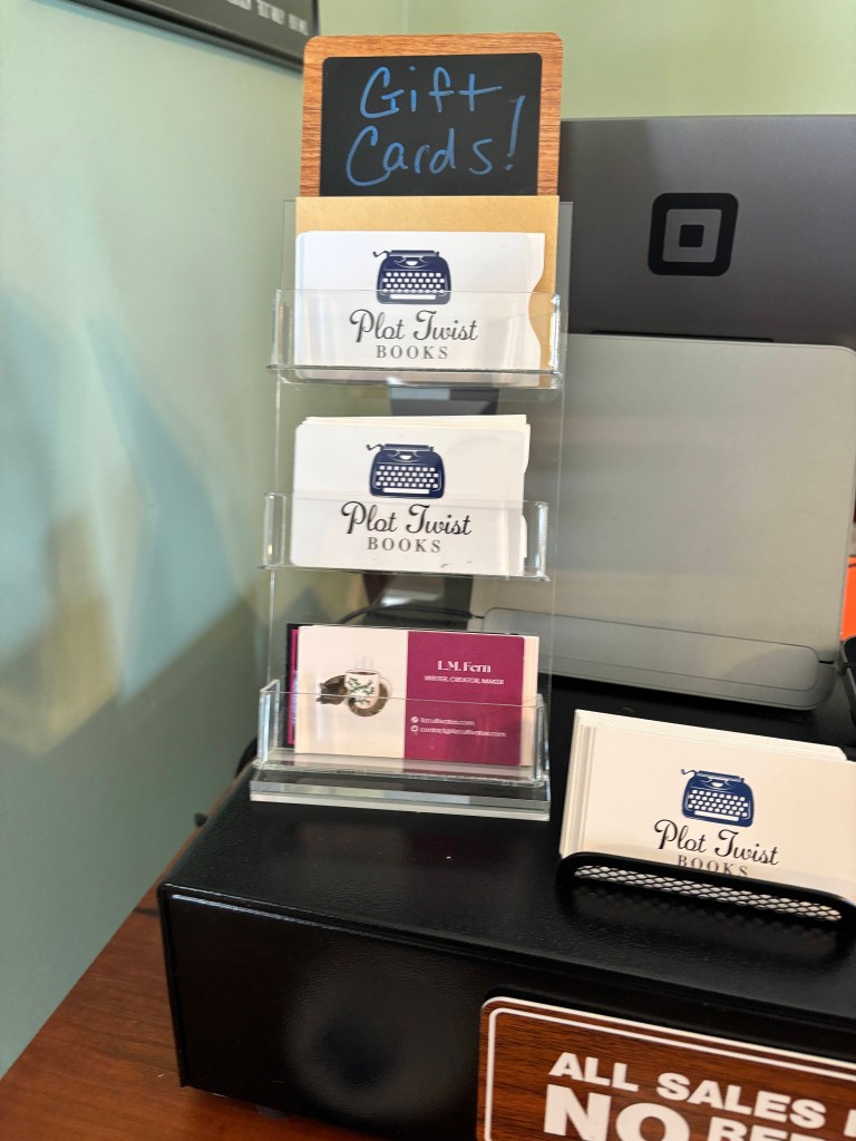 A stack of business cards and gift cards, with the bottom displaying the LM Fern business card, at Plot Twist Books